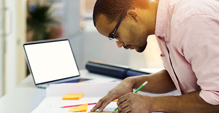 An architect stands at his desk with an open laptop beside him, drawing diagrams on drafting paper.