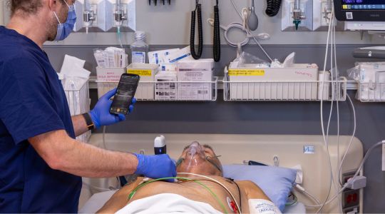 A nurse using a handheld ultrasound scanner on a man’s chest while observing the results on a phone, as he lays in a hospital bed.