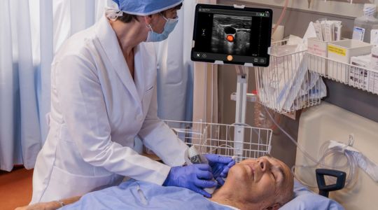 A nurse using a handheld ultrasound scanner on a man’s neck while observing the results on a tablet, as he lays in a hospital bed in a gown.