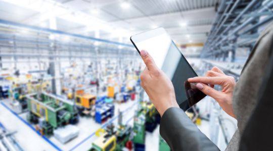 A businesswoman’s hands typing on a tablet inside a factory setting. 