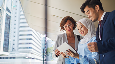 Une femme aux cheveux bouclés portant des lunettes, une femme portant un hijab blanc et un homme en complet bleu sont debout sur un balcon et regardent une tablette électronique.