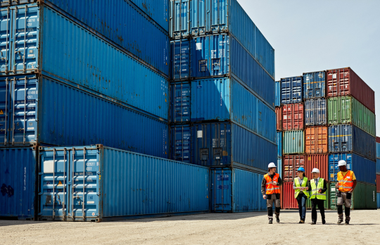 Un groupe de travailleurs portant des gilets de sécurité et des casques de protection se tient à côté d’une grande pile de conteneurs d’expédition