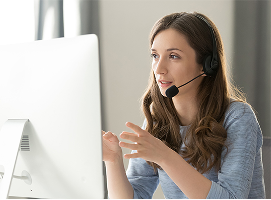 A woman facing a computer monitor speaks into a headset and gestures with her hands