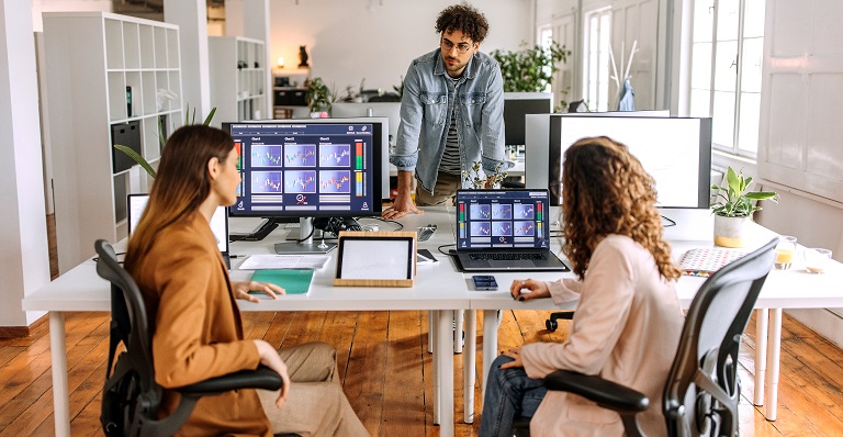 Three co-workers (one male and two females) look at colourful graphs on their computer screens.