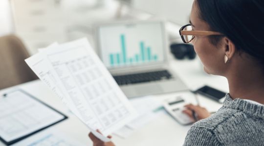 A woman with glasses looking at tax documents on paper and on her laptop.