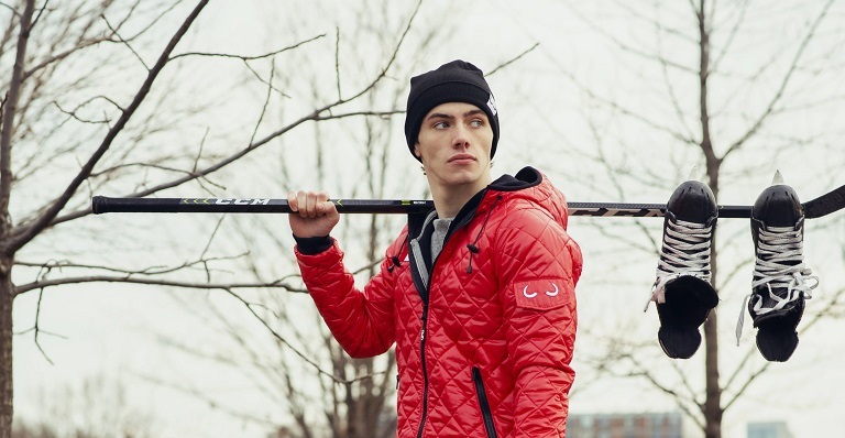 Man wearing Canada’s red Wuxly jacket carries hockey stick and skates.