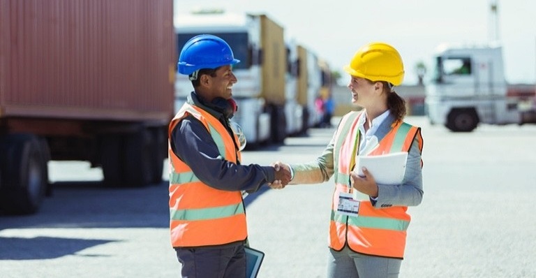 A man and a woman wearing construction hats shake hands and smile.