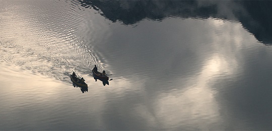 Deux canots, ayant chacun à son bord deux personnes, glissent côte à côte sur des eaux calmes.