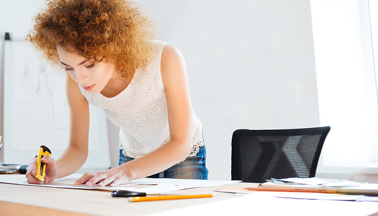A small business owner in a sunlit office stands at her desk measuring and scoring a sheet of paper.